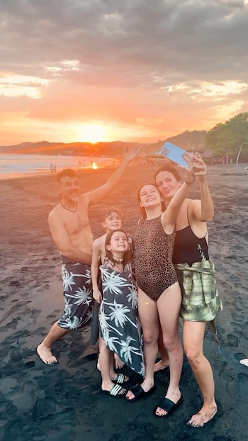 Cinco personas, dos adultos y tres niños, se toman una selfi en una playa de arena oscura con el mar y un atardecer anaranjado de fondo