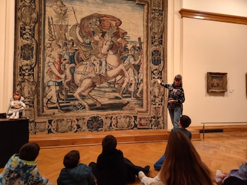 Vista interior de un museo con una guía señalando un gran tapiz militar a niños sentados en el suelo. A la izquierda, una muñeca de porcelana en un pedestal