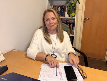 Mujer rubia de mediana edad sentada en un escritorio de madera. Viste suéter blanco. Sobre la mesa hay una agenda, bolígrafo, móvil y gafas