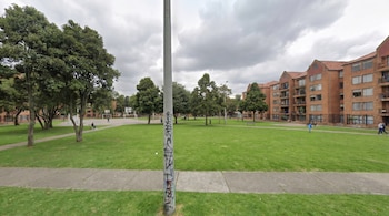 Vista panorámica de un extenso parque urbano con césped verde, árboles, un poste de luz con grafitis, una cancha de baloncesto y edificios de ladrillo bajo un cielo nublado.