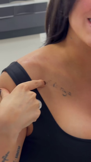 Una persona señala con el dedo un tatuaje de tinta negra con trazo tenue en la clavícula de una mujer