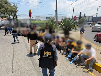 Agentes del Ministerio Público de Guatemala con chalecos "MP" están en una acera, supervisando a personas sentadas. Se ven edificios, vehículos y un letrero de McDonald's