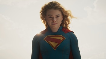 Primer plano de Milly Alcock como Supergirl con el traje azul y rojo, cabello rubio al viento, mirando directamente a la cámara bajo un cielo luminoso