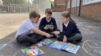 Tres niños, dos niños y una niña, sentados en el suelo de un patio escolar, intercambian figuritas del Mundial FIFA 2026 y miran un álbum.