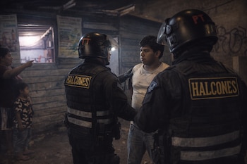 Dos agentes de policía con uniformes tácticos y cascos, uno con luz frontal, interactúan con un hombre joven. Al fondo, una mujer señala y un niño observa.