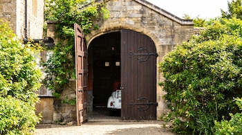 Un coche clásico blanco con capota roja se ve parcialmente dentro de un garaje de piedra con arco, puertas de madera abiertas y vegetación exuberante a los lados.