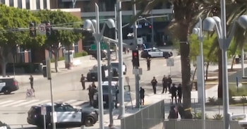 Una calle urbana con varios coches de policía y numerosos oficiales. Se observan semáforos, árboles y edificios al fondo, con cinta amarilla marcando un área