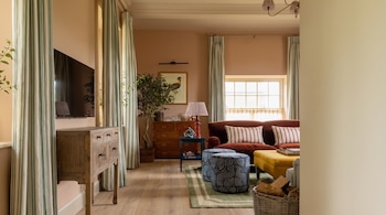 Sala com sofá, pufe, duas cadeiras, mesa de centro, móveis de madeira, planta, cortina curva, janela, quadro, abajur, cesto de lenha.