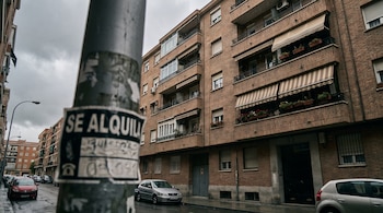 Poste metálico con cartel "Se alquila" desenfocado. Edificio residencial de ladrillo con balcones y toldos. Coches estacionados en calle mojada bajo cielo gris.