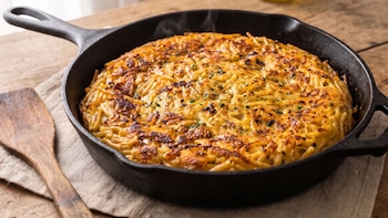 Sartén de hierro fundido con tortilla de fideos dorada, junto a una espátula y un paño sobre una mesa de madera