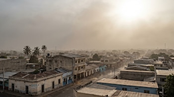 Vista aérea de una ciudad salvadoreña con calles, edificios y palmeras bajo un cielo opaco y una densa bruma grisácea. El sol brilla difuso en el horizonte.