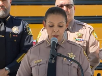 Oficial femenina en uniforme marrón de seguridad hablando a un micrófono, con otros agentes y un autobús escolar amarillo detrás