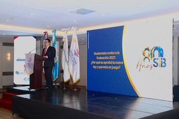 Un hombre con traje y corbata habla en un atril sobre un escenario. Detrás hay banderas y una pantalla con el texto 'Guatemala rumbo a la Evaluación 2027' y el logo '80 Años SIB'