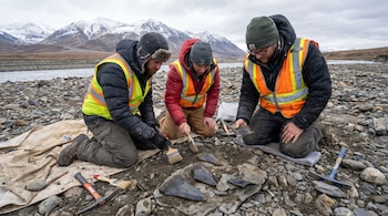 Tres paleontólogos agachados examinando restos fósiles de dientes grandes en un terreno rocoso, cerca de un río, con montañas nevadas y cielo nublado de fondo en Alaska.