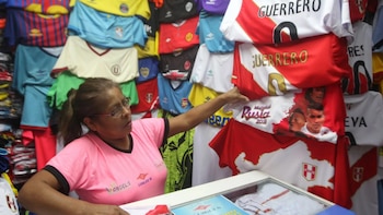Mujer con lentes y camiseta rosa señala una camiseta de fútbol blanca y roja con el logo de Perú; detrás, una pared llena de camisetas deportivas de varios equipos y colores