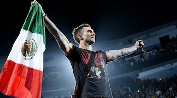 Adam Levine, vocalista de Maroon 5, con tatuajes en los brazos, levanta la bandera mexicana con ambos brazos extendidos en un escenario de concierto oscuro.