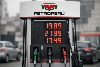 Primer plano de un surtidor de gasolina de Petroperú con precios digitales rojos y altos en pantalla, y boquillas de colores verde, negro y rojo. Autos desenfocados al fondo.