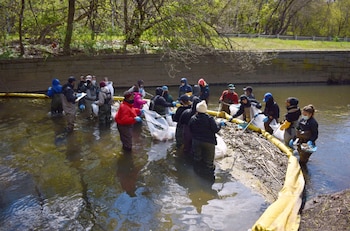 Grupo de personas con botas de vadeo, guantes y mascarillas recogen desechos en un río. Usan bolsas, pinzas y un flotador amarillo. Árboles verdes y muro de piedra visibles