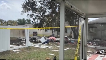 Escombros de un avión esparcidos en un patio trasero cercado. Se observa cinta amarilla de "SHERIFF'S LINE DO NOT CROSS" y marcas de quemaduras en el suelo