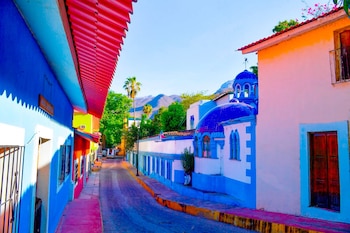 Una calle adoquinada bordea casas de múltiples colores, dos edificios con cúpulas azules, palmeras y montañas al fondo