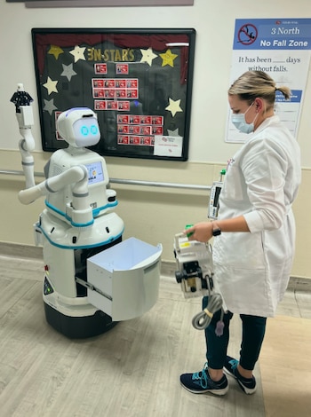 Un robot de entrega blanco y azul con detalles celestes y un cajón abierto, llamado Moxi, se encuentra frente a una enfermera en un pasillo de hospital