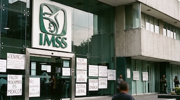 Edificio del IMSS con logo verde, entrada de cristal y carteles de denuncia pegados, con figuras borrosas de personas cerca de la entrada.