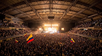 Pabellón cubierto con gradas y pista repletas de público que ondea banderas de Colombia frente a un escenario iluminado.