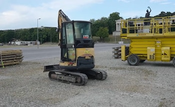 Excavadora compacta amarilla, plataforma elevadora amarilla, pilas de madera, losas apiladas, terreno de grava, árboles y edificaciones al fondo