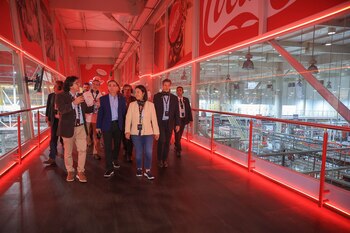 Un grupo de seis personas camina sobre una pasarela iluminada de rojo dentro de una fábrica, con el logotipo de Coca-Cola en la pared y maquinaria al fondo