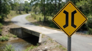 Señal de tránsito amarilla con símbolo de puente angosto en primer plano; detrás, una carretera rural curva, un pequeño puente y un arroyo rodeado de árboles.