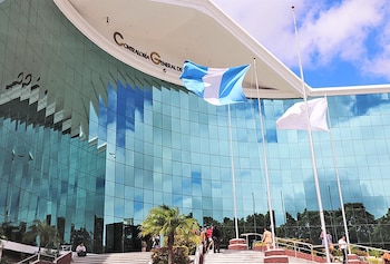 Fachada curva de cristal de la Contraloría General de Cuentas de Guatemala con bandera de Guatemala ondeando, otras banderas y personas en las escaleras