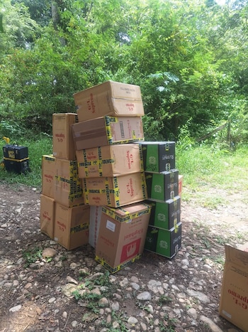 Pilas de cajas de cartón de cigarrillos G*L Supremo, Elephant y Richman, con cinta de Evidencia MP, en camino de tierra con vegetación