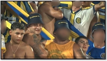 Primer plano de un grupo de hombres en una tribuna. Varios visten camisetas del club o gorros azules con la inscripción 'La Guerrilla'. Hay banderas azules y amarillas