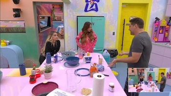 Tres personas en una cocina colorida con mesa rosada. Hay tazones, botellas, jarra, servilleta y alimentos. Pantalla muestra la misma escena