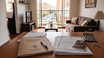 Documentos de compraventa de apartamento, pasaporte, llaves y calculadora sobre una mesa en una sala con sofá y balcón, vista a edificios.