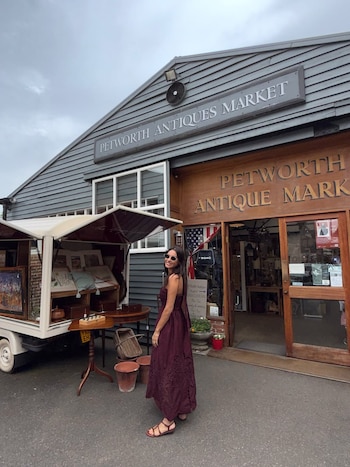 Pampita sonríe con gafas de sol y un vestido largo borgoña. Está de pie frente a la entrada de madera de un mercado de antigüedades y un puesto exterior