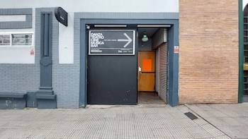 Vista exterior de la entrada a Planta Inclán, un edificio con paredes de ladrillo gris y detalles en azul oscuro, destacando un letrero negro y una puerta de madera