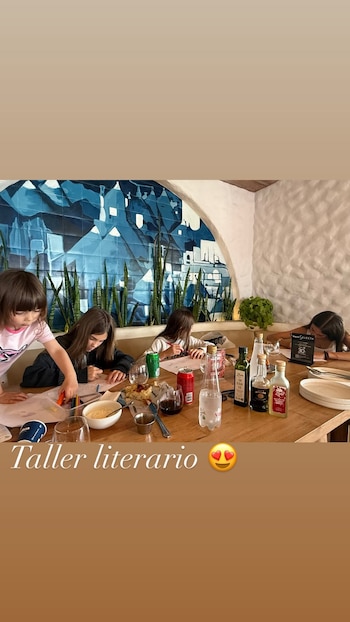 Cuatro niñas escriben sobre hojas en una mesa de madera con vasos, botellas y un texto superpuesto que dice Taller literario