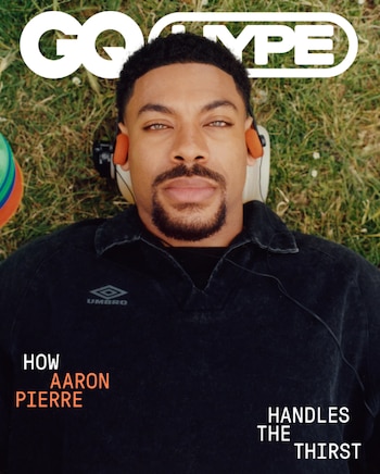Hombre joven con barba recostado en césped, con ojos claros, camiseta Umbro oscura y auriculares naranja. Arriba, logo GQ Type y texto de revista