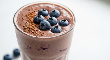 Primer plano de un batido cremoso de color marrón en un vaso, con arándanos flotando en la superficie y una pizca de cacao en polvo espolvoreado.