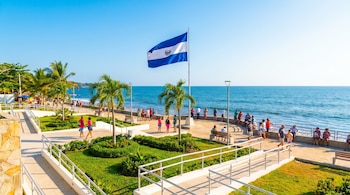 Vista panorámica del Malecón del Puerto de La Libertad en El Salvador, con la bandera nacional, turistas, palmeras y áreas verdes junto al mar.