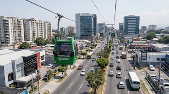 Vista aérea de cabinas verdes del Aerometro sobre una avenida urbana con tráfico, rodeada de edificios altos y residenciales bajo un cielo despejado.