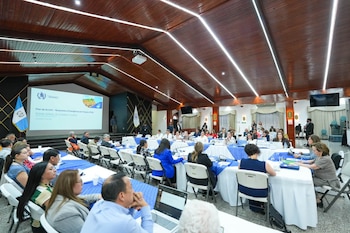 Personas sentadas a lo largo de mesas con manteles blancos y azules en un salón con techo de madera y la bandera de Guatemala al fondo