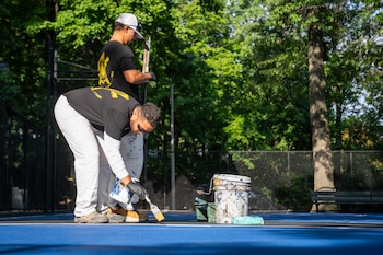 Dos trabajadores con vestimenta de obra aplican pintura en el suelo de una cancha azul al aire libre rodeada de árboles