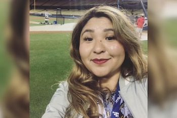 Una mujer sonriente con cabello castaño claro y labial rojo se toma una fotografía en un campo de béisbol