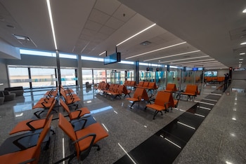 Filas de sillas naranjas en una sala de espera vacía con piso brillante, luces lineales en el techo y ventanales al fondo