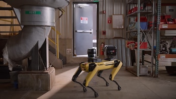 Robot amarillo y negro de cuatro patas Boston Dynamics Spot con sensores en el lomo, en un almacén industrial con estanterías y tuberías