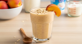 Primer plano de un vaso de licuado de durazno, avena y yogur con una rodaja de durazno y avena encima, junto a una cuchara de madera sobre una mesa clara.