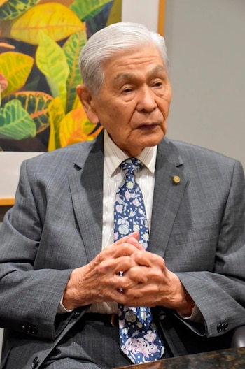 George Ryoichi Ariyoshi, un hombre mayor con traje gris y corbata estampada, sentado con las manos entrelazadas. De fondo, una pintura abstracta de hojas