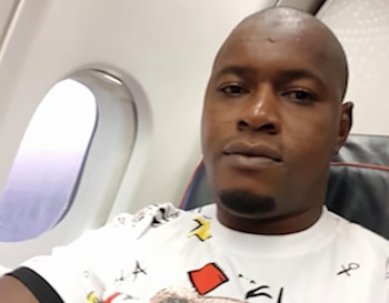 Hombre de cabeza rapada y perilla con camiseta blanca estampada, sentado junto a la ventanilla de un avión